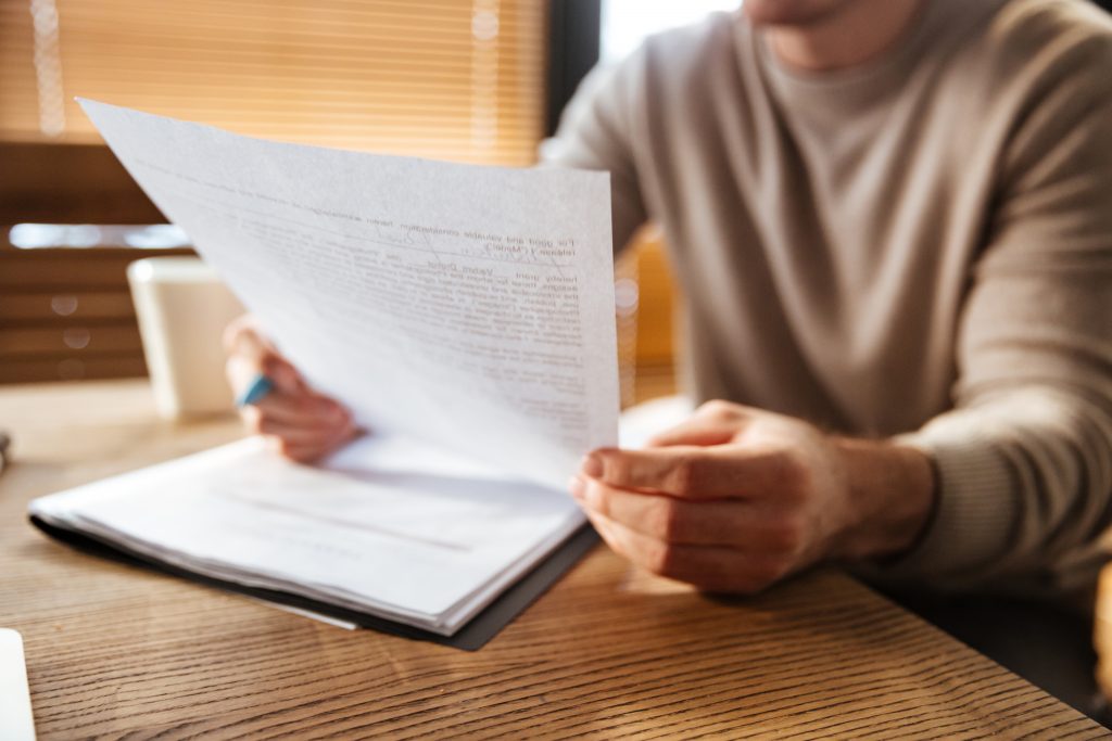 cropped photo of young man reviewing a document in one hand and holing a pen in the other on top of more documents