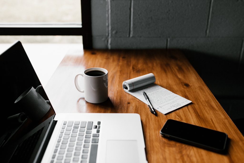 a laptop, a pen and notepad, a coffee mug, and a phone on top of a wooden desk