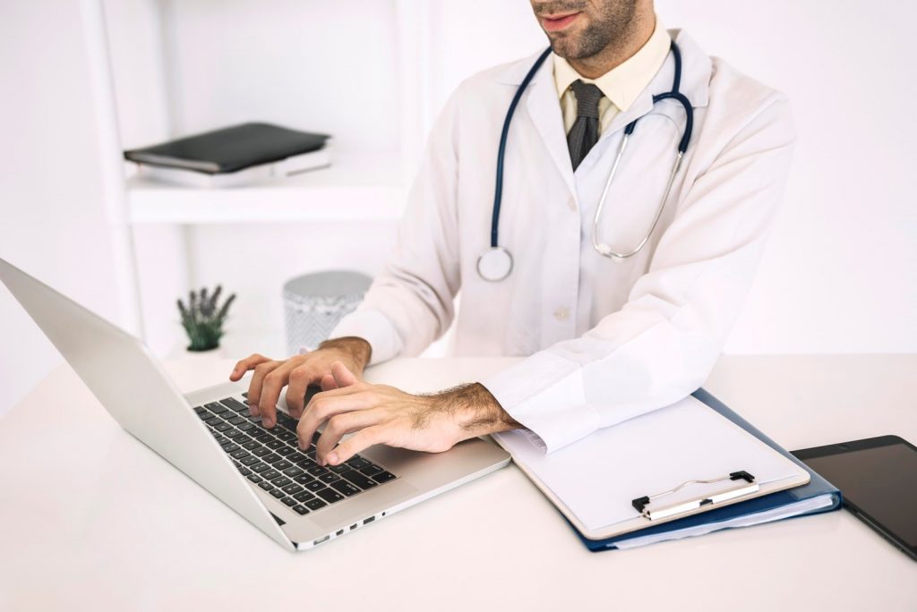male medical professional typing medical transcriptions on a laptop beside documents on a clipboard