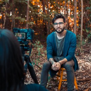 a male subject is being interviewed in a forest by a female interviewer with DSLR camera and tripod for a documentary interview